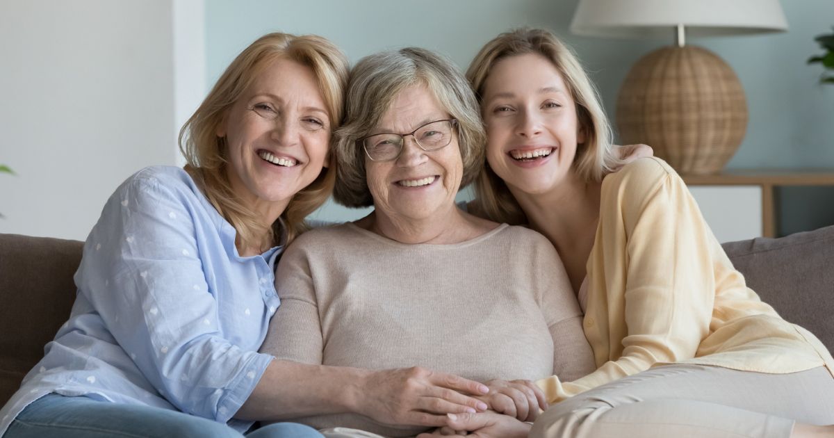 Three generations of women sitting together on a couch, smiling and showing family connection and support for aging parents.