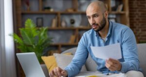 Man reviewing a financial document at his laptop while comparing investment tax details at home.