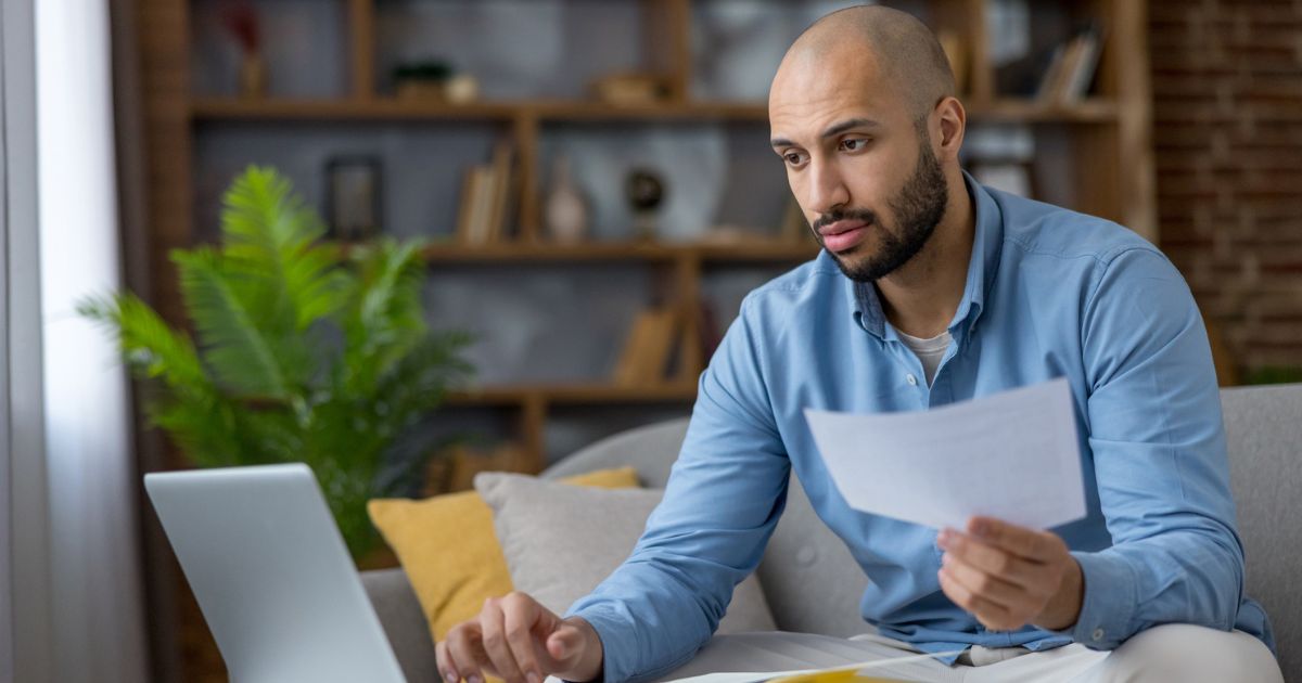 Man reviewing a financial document at his laptop while comparing investment tax details at home.